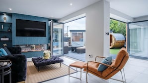 A modern living room with a flat-screen TV, comfortable seating, and a glass door leading to an outdoor area with a wooden barrel.