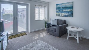 A modern living room with a grey sofa, a white round table, and a painting of a seascape. - Sea Breeze Apt 1 - 1 Bedroom - Horton Beach (Swansea)