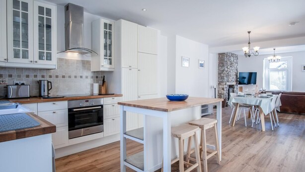 Cocina moderna con armarios blancos, horno integrado y encimera de madera. Hay una zona de comedor con una mesa y sillas, y una pared de piedra al fondo.