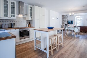Cocina moderna con armarios blancos, horno integrado y encimera de madera. Hay una zona de comedor con una mesa y sillas, y una pared de piedra al fondo.