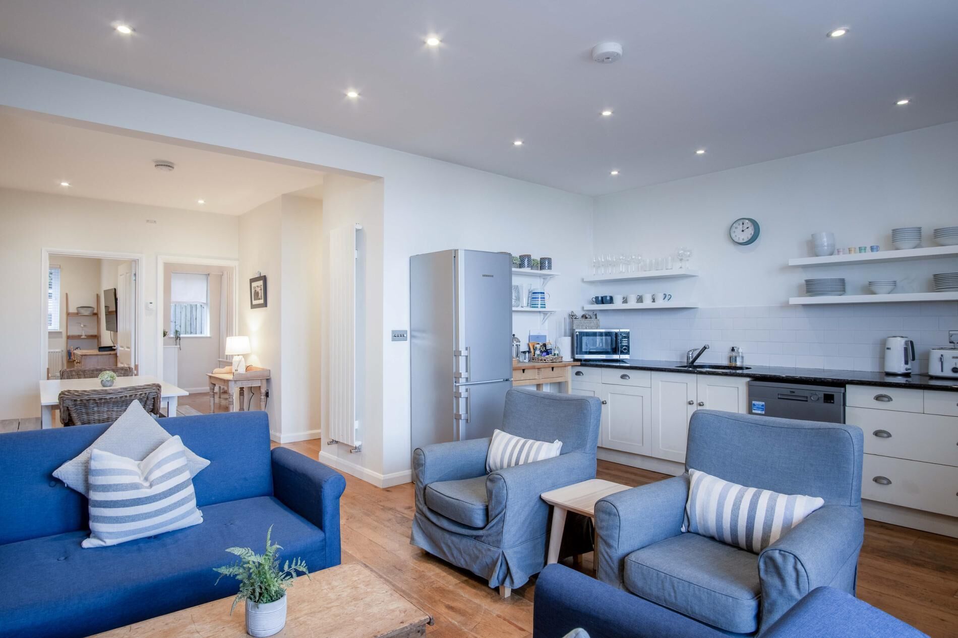 Un salon moderne avec un canapé et des fauteuils bleus, un petit coin repas et une cuisine dotée de meubles blancs et d’un four à micro-ondes.