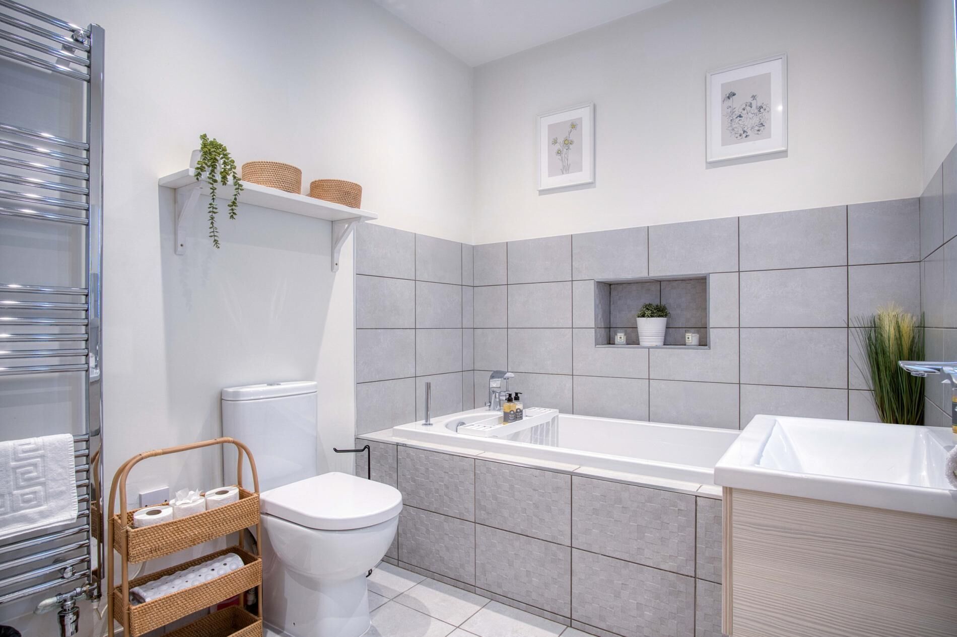 A modern bathroom with a white toilet, a freestanding bathtub, and a shower area. There are two framed pictures on the wall, a small shelf with toiletries, and a wicker basket with towels.