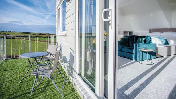 A modern hotel room with a glass door leading to a balcony. The room features a bed, a sofa, and a small table. The balcony has a table and two chairs. - The Penthouse - 1 Bedroom Apartment - Ocean Breeze (Swansea)