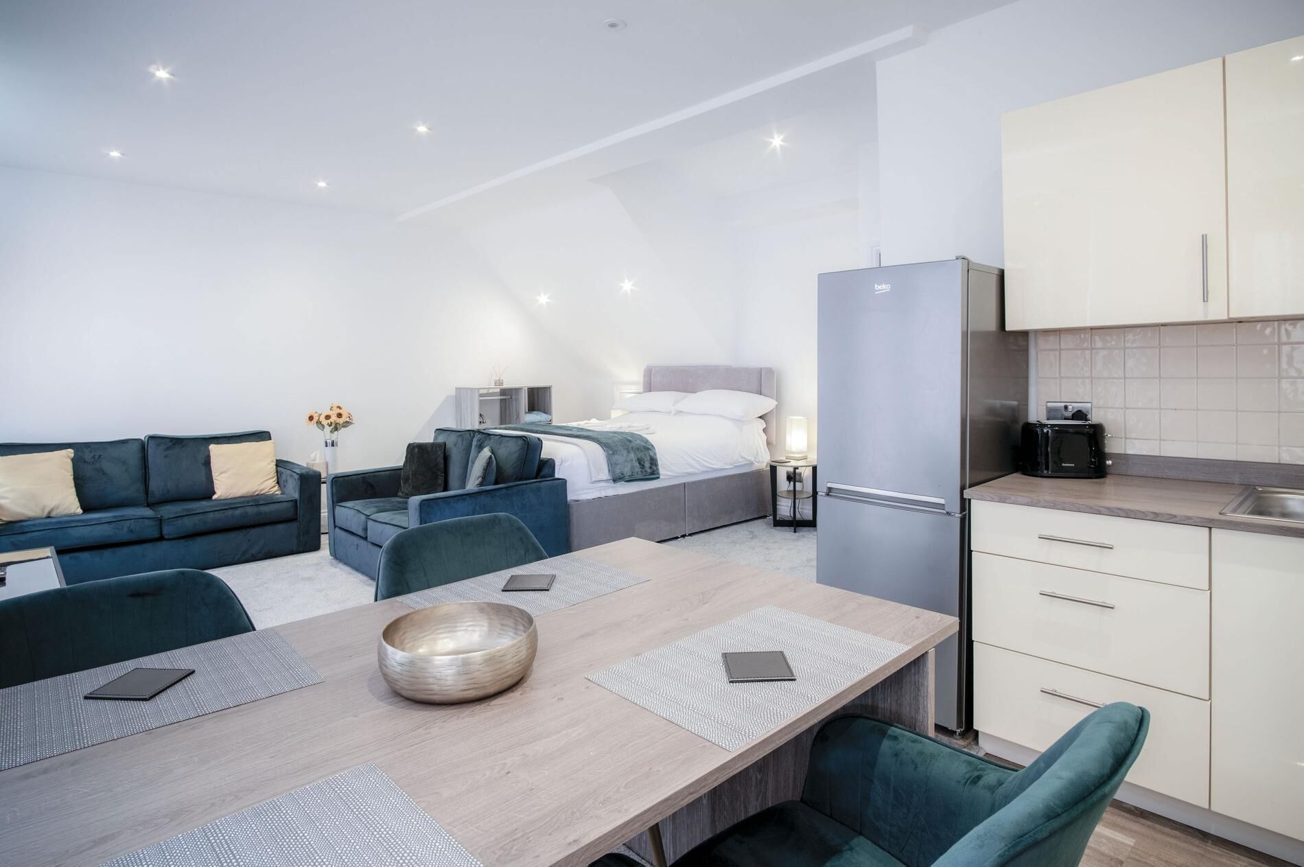 A modern, well-lit room with a bed, sofa, dining table, and kitchenette.