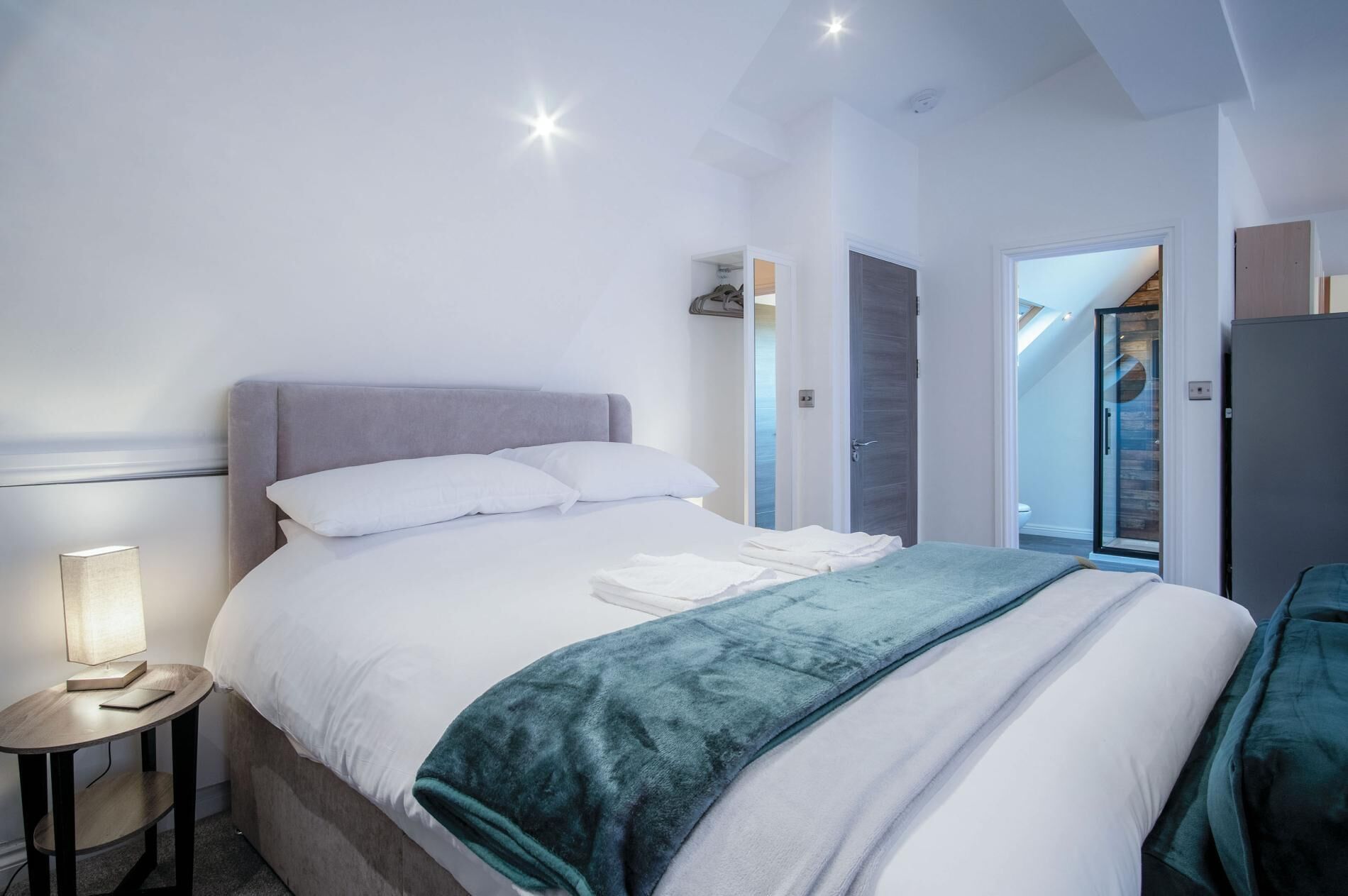 A neatly made bed with white linens and a teal throw blanket, a bedside table with a lamp, and a door leading to a bathroom.