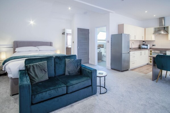 A modern bedroom with a bed, a blue sofa, and a small table. There is a kitchen area with a refrigerator and a microwave. - The Penthouse - 1 Bedroom Apartment - Ocean Breeze (Swansea)