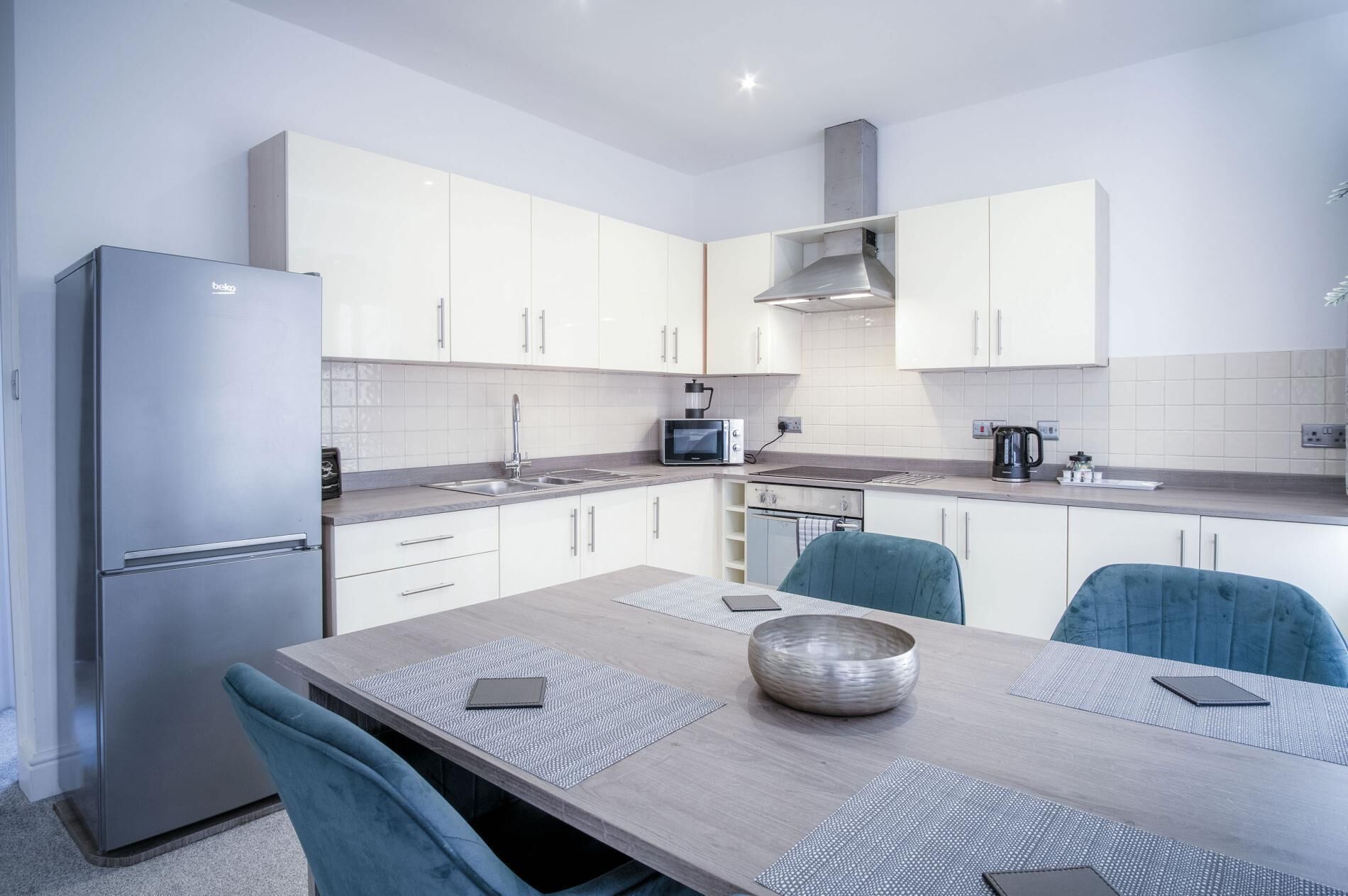 A modern kitchen with white cabinets, a stainless steel refrigerator, a microwave, and a gas stove. There is a dining table with blue chairs and a large bowl on it.