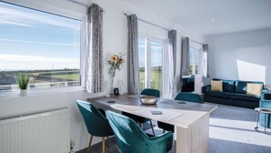 A modern living room with a dining area, large windows, and a view of the countryside. - The Penthouse - 1 Bedroom Apartment - Ocean Breeze (Swansea)