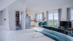 A modern hotel room with a large bed, a TV, a dining area with teal chairs, and a view of the sea. - The Penthouse - 1 Bedroom Apartment - Ocean Breeze (Swansea)