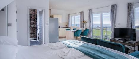 Ett modernt hotellrum med en stor säng, en TV, en matplats med turkosa stolar och utsikt över havet.