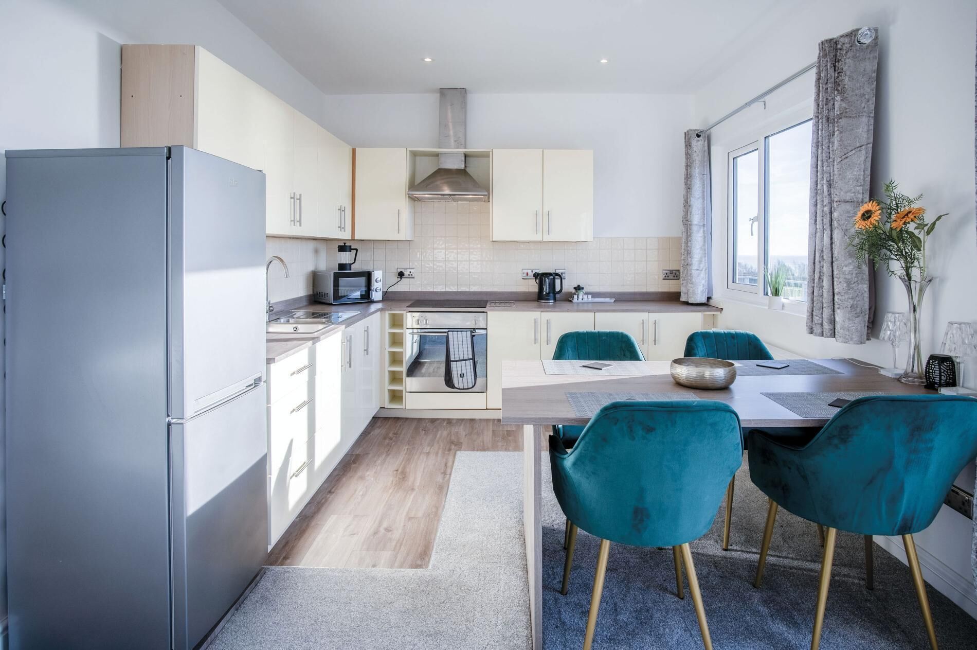 Una cocina moderna con armarios blancos, un refrigerador de acero inoxidable, un microondas y una cafetera. Cuenta con una zona de comedor con una mesa de madera y sillas color turquesa.