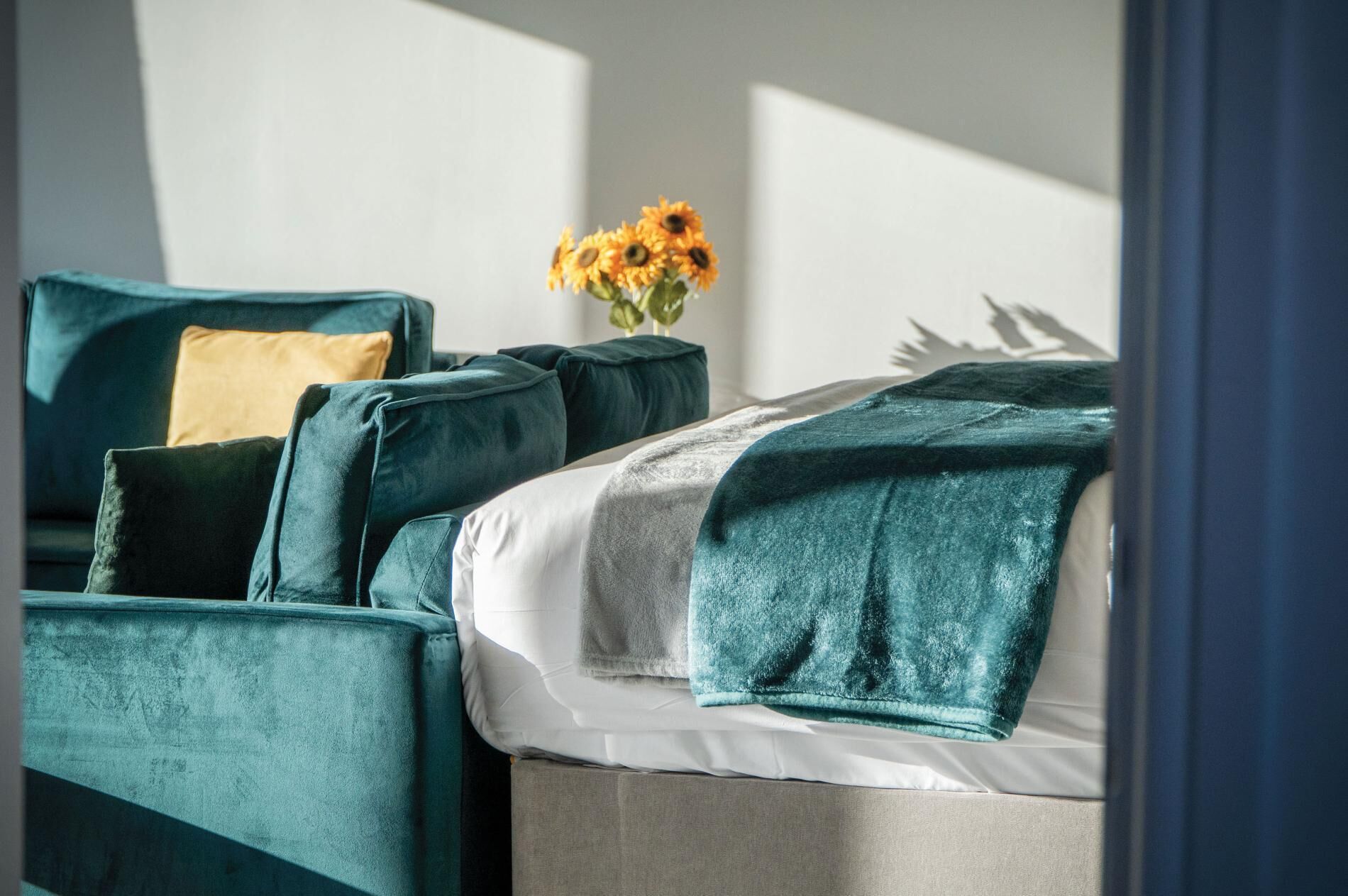 A modern room with a teal upholstered sofa, a white bed with a teal throw blanket, and a vase with yellow flowers on a side table.