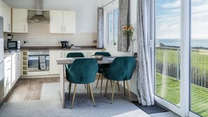 A modern kitchen with white cabinets, a built-in oven, and a dining area with teal chairs and a wooden table. - The Penthouse - 1 Bedroom Apartment - Ocean Breeze (Swansea)