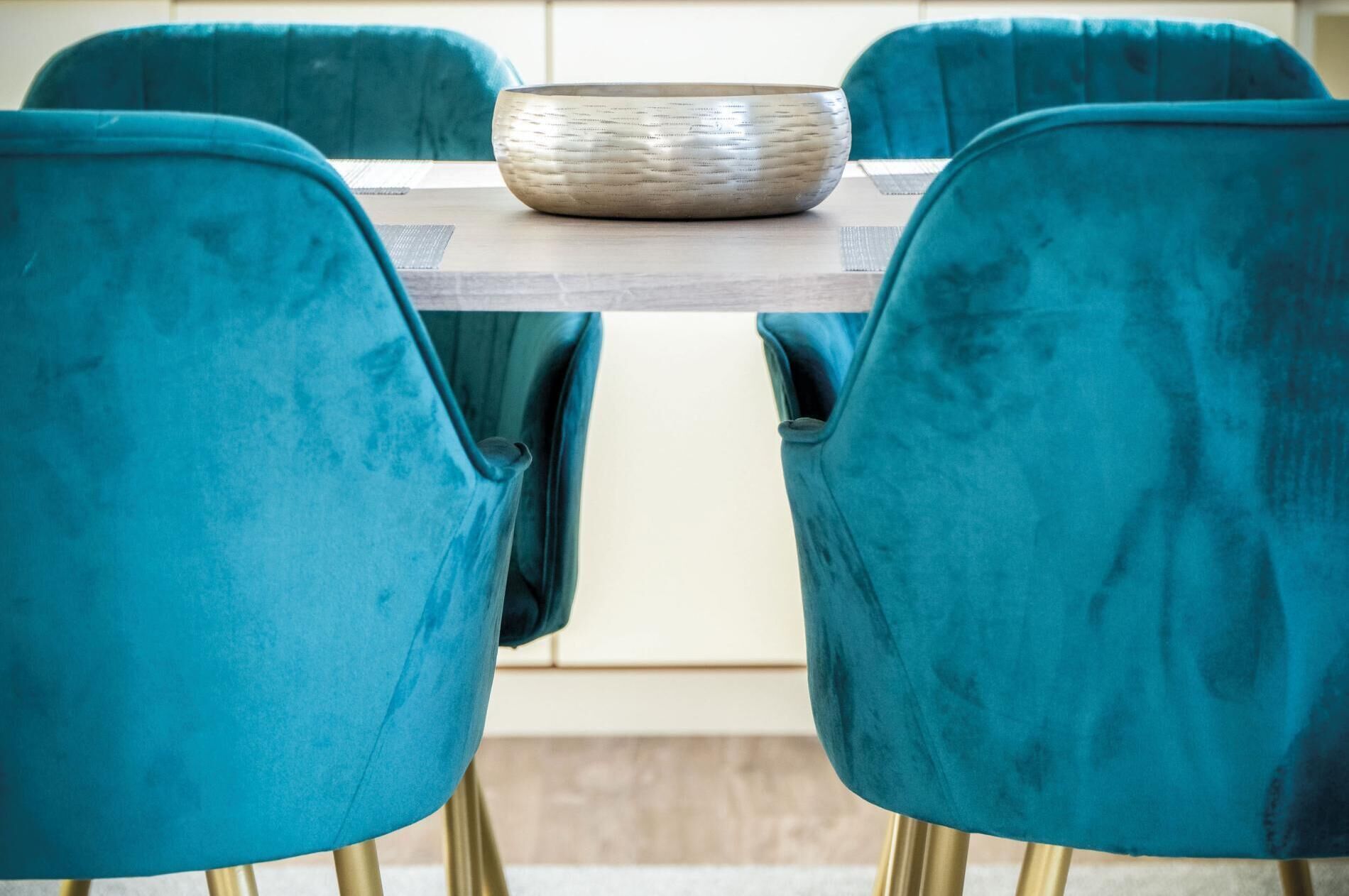 A dining table with a woven bowl centerpiece, surrounded by four teal upholstered chairs with gold legs.