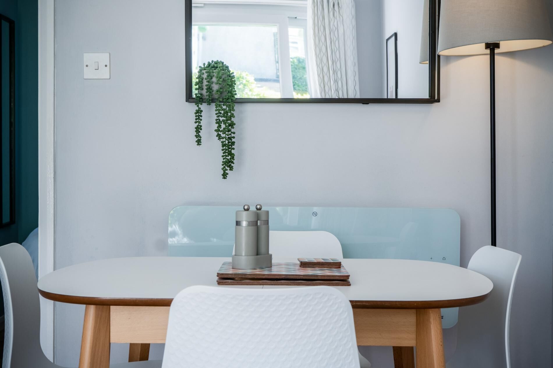 Ein moderner Essbereich mit einem weißen Tisch, Holzstühlen, einer hellblauen Bank, einer kleinen Pflanze, einer Kaffeemaschine und einer Lampe.