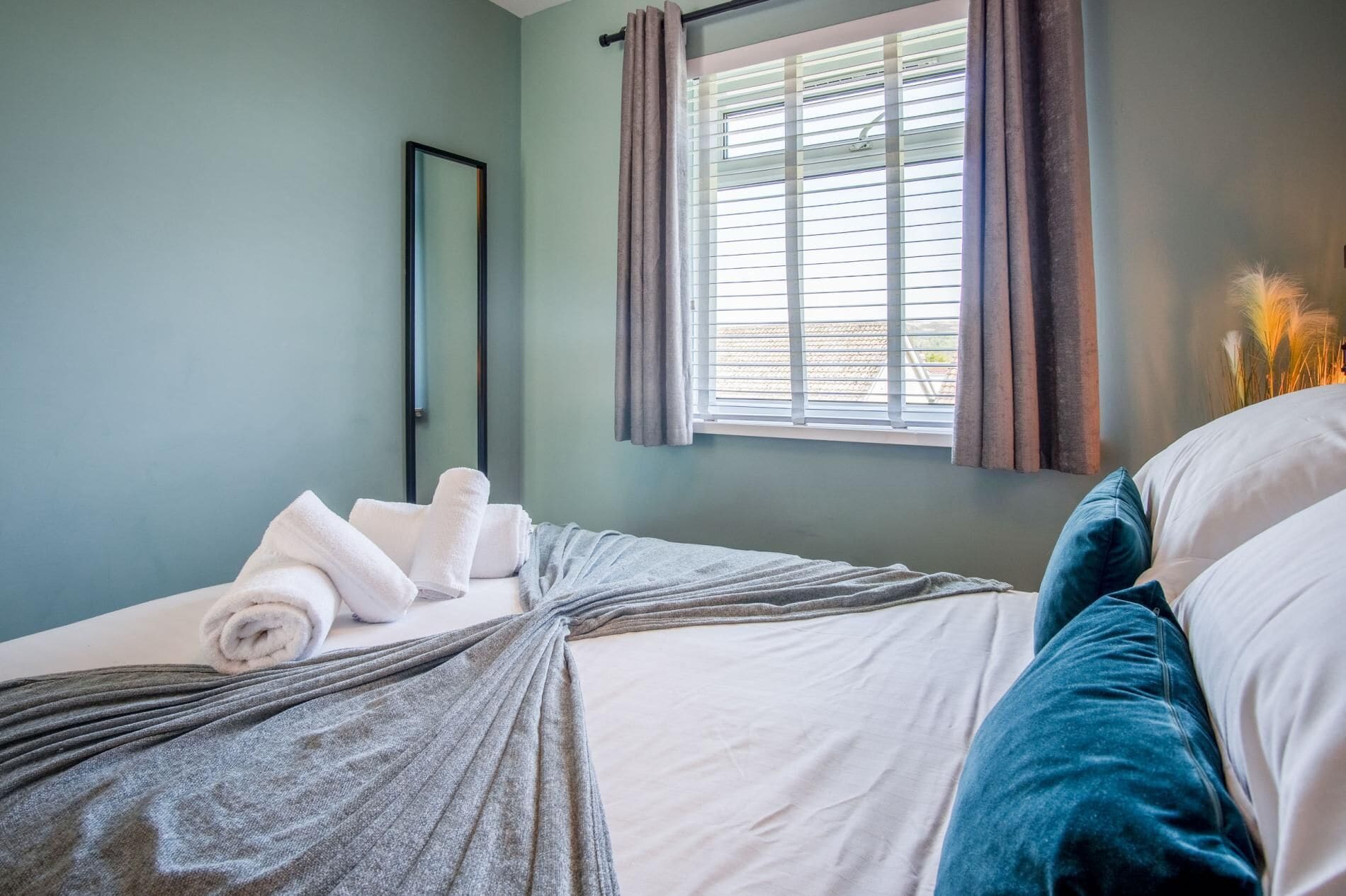A neatly made bed with white and grey linens, a window with blinds, and a mirror on the wall.
