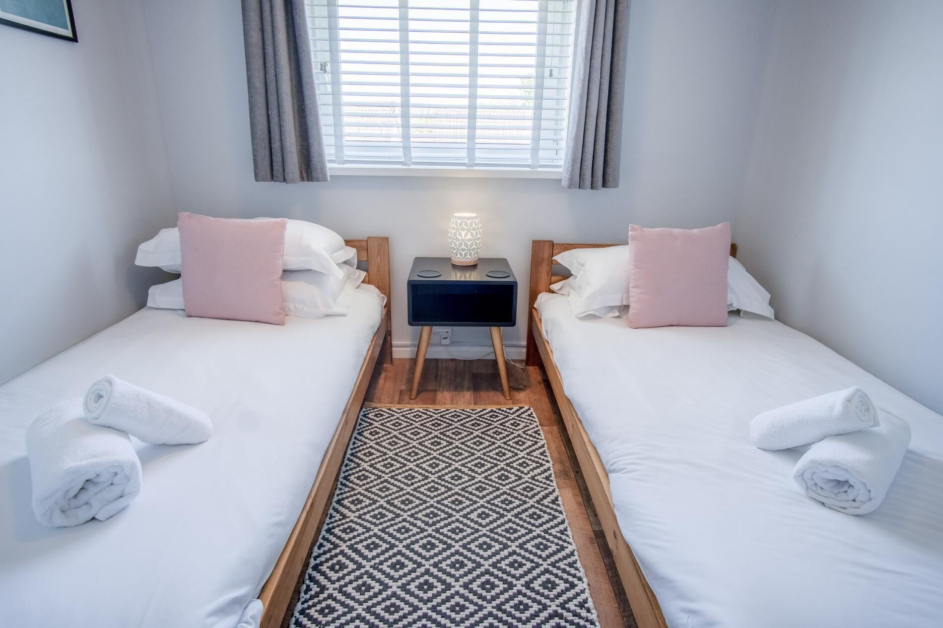 A hotel room with two single beds, a nightstand with a lamp, a patterned rug, and a window with curtains.