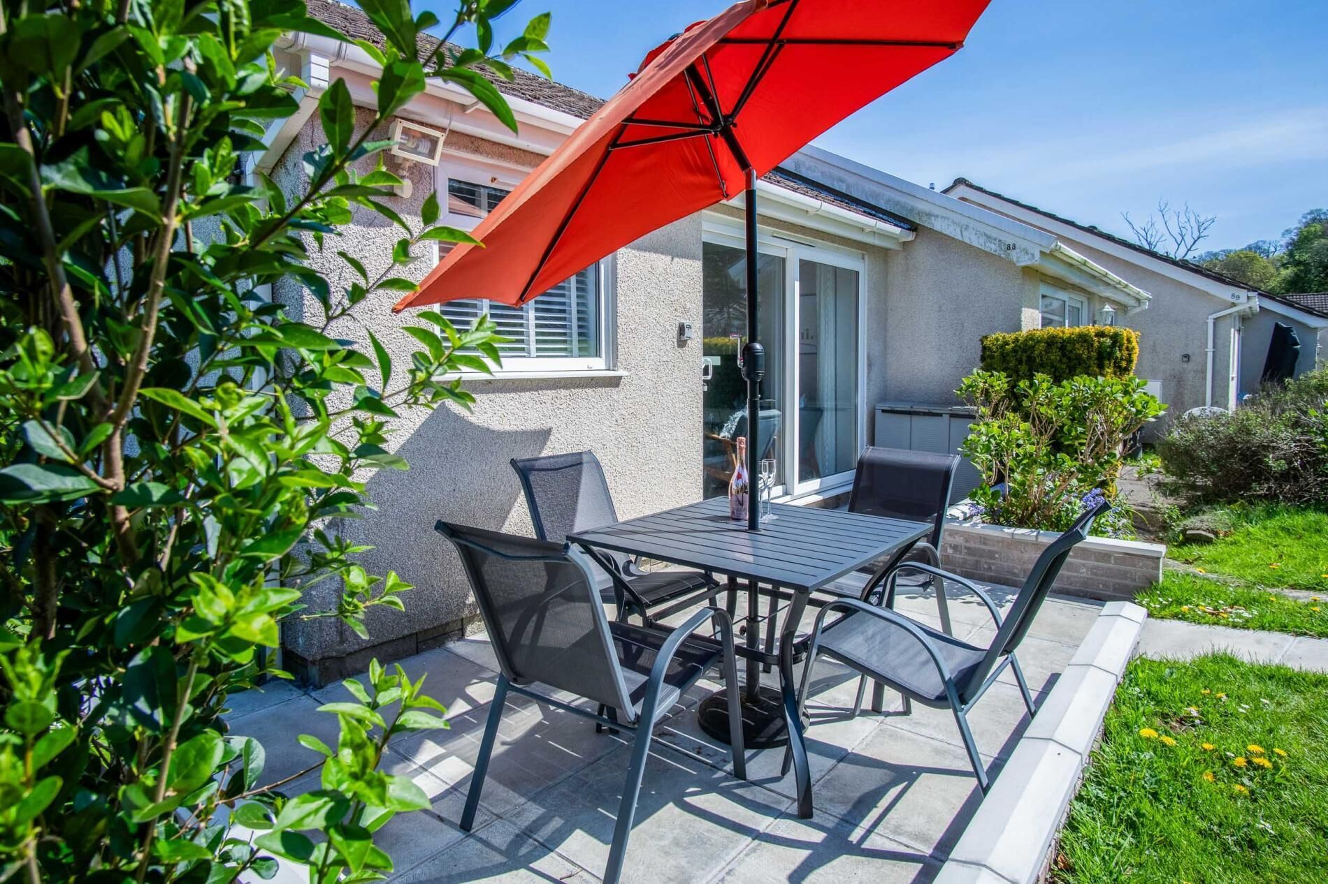 Une terrasse extérieure avec une table et des chaises, un parasol rouge et une maison en arrière-plan.