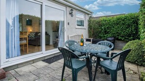 A patio with a table and chairs, a bottle of champagne, and a view into the house through glass doors. - Bayview 58 - 2 Bedroom Chalet - Bayview Oxwich (Swansea)