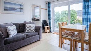 A modern living room with a grey sofa, a wooden dining table with chairs, a white coffee table, and a large window with blue curtains. - Bayview 58 - 2 Bedroom Chalet - Bayview Oxwich (Swansea)