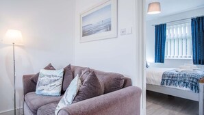 A modern room with a grey sofa, a floor lamp, a bed with a blue and white blanket, and a window with blue curtains. - Bayview 58 - 2 Bedroom Chalet - Bayview Oxwich (Swansea)