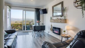 A modern living room with a sofa, a dining table with chairs, a TV, and a fireplace. - Bayview 32 - 2 Bedroom Chalet - Bayview Oxwich (Swansea)