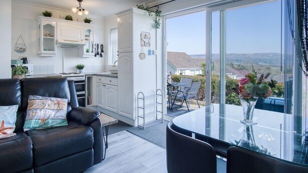 A compact kitchen with white cabinets, a dining area with a glass table and chairs, and a view of a hilly landscape through large windows. - Bayview 32 - 2 Bedroom Chalet - Bayview Oxwich (Swansea)