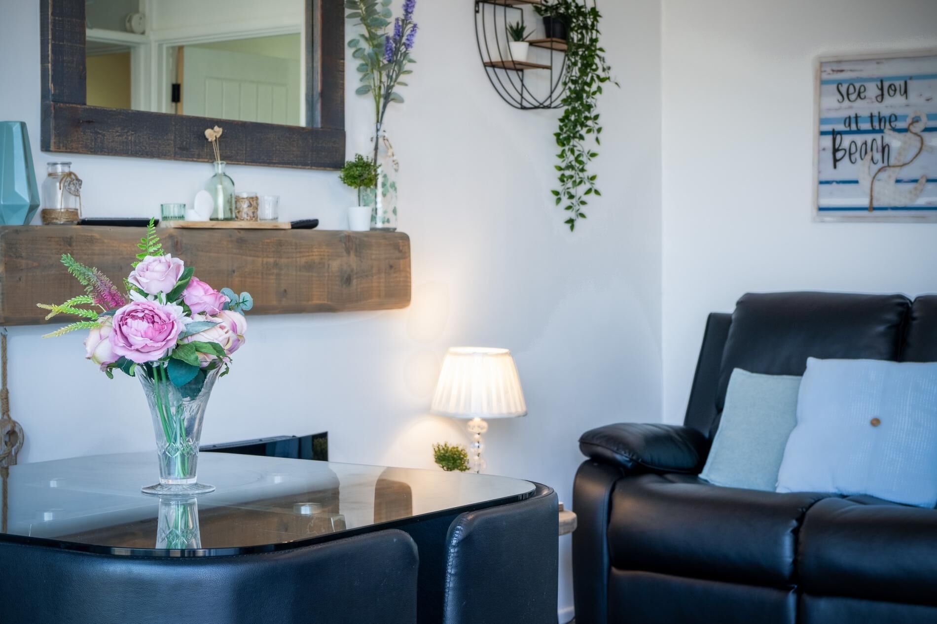 A living room with a glass coffee table, a black leather sofa, a vase with flowers, a lamp, and a wall-mounted shelf with plants.