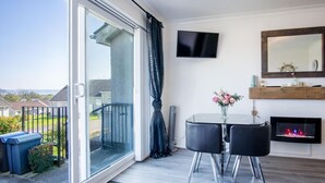 A balcony with a sliding glass door, a small table with two chairs, a flat-screen TV mounted on the wall, and a fireplace. - Bayview 32 - 2 Bedroom Chalet - Bayview Oxwich (Swansea)