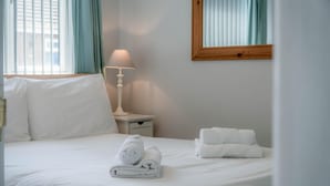 Ein ordentlich bezogenes Bett mit weißen Leinentüchern, ein aufgerolltes Handtuch und ein gefaltetes Handtuch auf einem weißen Nachttisch daneben.