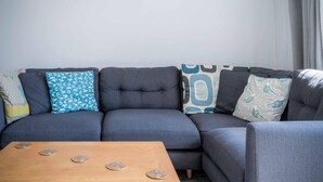 Ein graues Ecksofa mit vielen Kissen, ein Couchtisch aus Holz mit dekorativen Untersetzern und gerahmte Poster an der Wand.