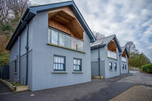 A modern two-story house with a grey exterior, large windows, and a wooden balcony. - Ty Megan - 3 Bedroom Cottage - Bayview Oxwich (Swansea)