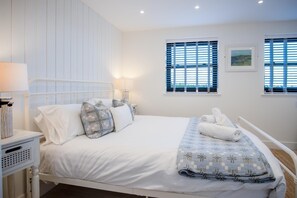 A neatly made bed with white linens and patterned pillows, a bedside table with a lamp, and two windows with blinds. - Ty Megan - 3 Bedroom Cottage - Bayview Oxwich (Swansea)