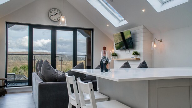A modern kitchen with a large island, a dining area with a sofa and chairs, a TV mounted on the wall, and a view of the countryside through large windows.