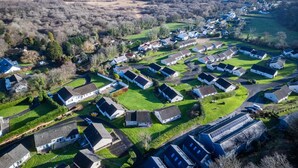 An aerial view of a residential area with houses arranged in a grid pattern, surrounded by green fields and a coastline in the background.