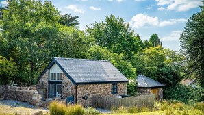 A stone cottage with a sloped roof, surrounded by lush greenery and trees.