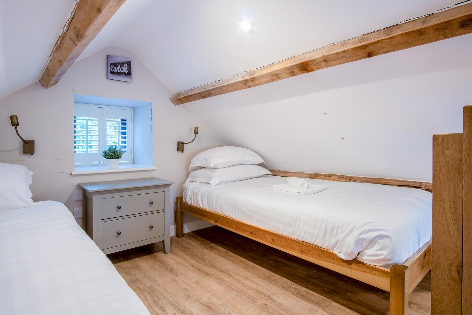 A bedroom with a wooden bed, a nightstand, a window with blinds, and a wall-mounted lamp.