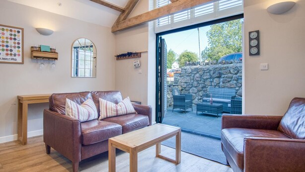 A cozy living room with a brown leather sofa, a wooden coffee table, and wicker chairs. A stone wall and a car are visible outside.