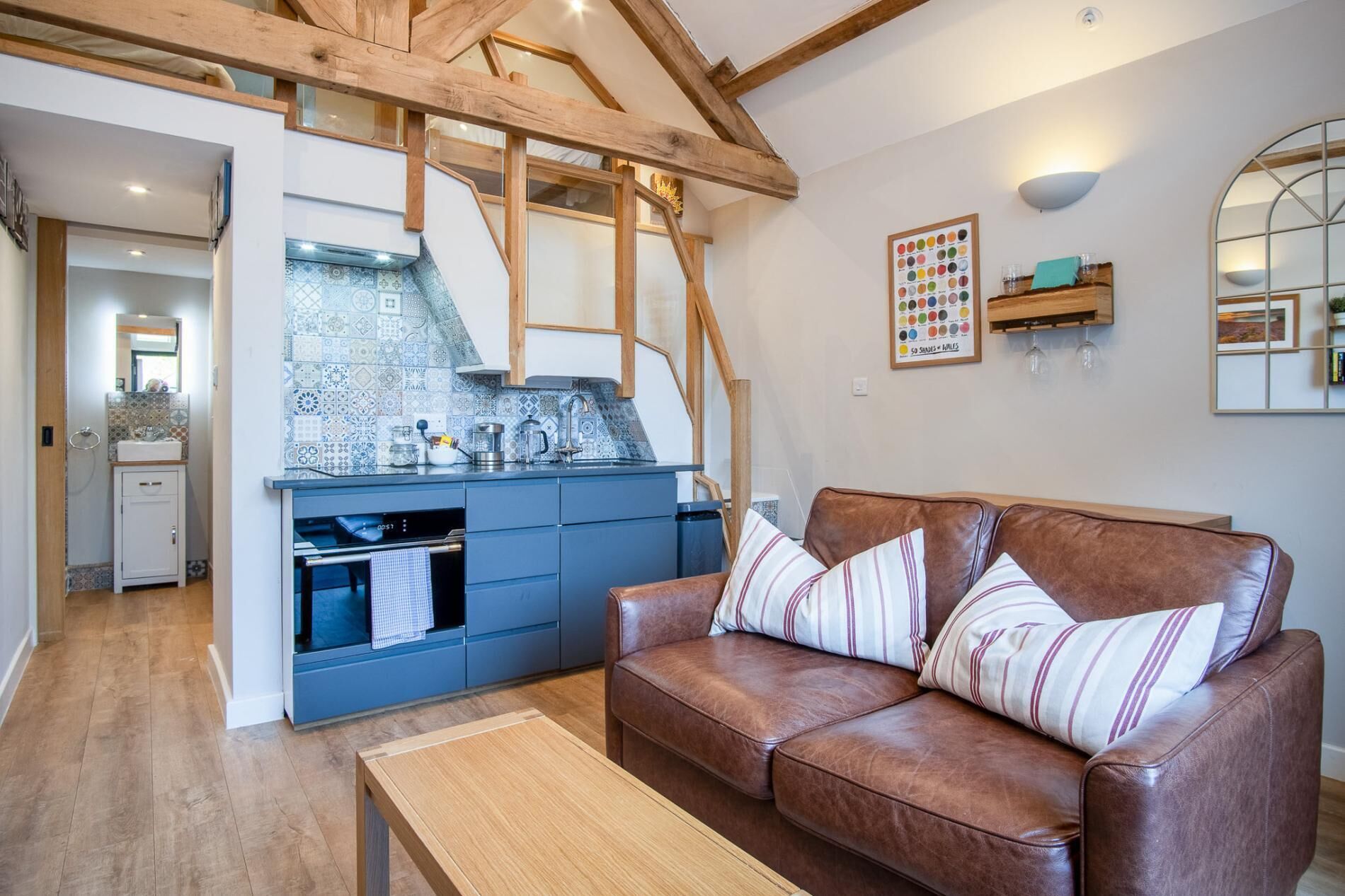 A cozy living room with a brown leather sofa, a wooden coffee table, and a kitchen area with blue cabinets and a tiled backsplash.