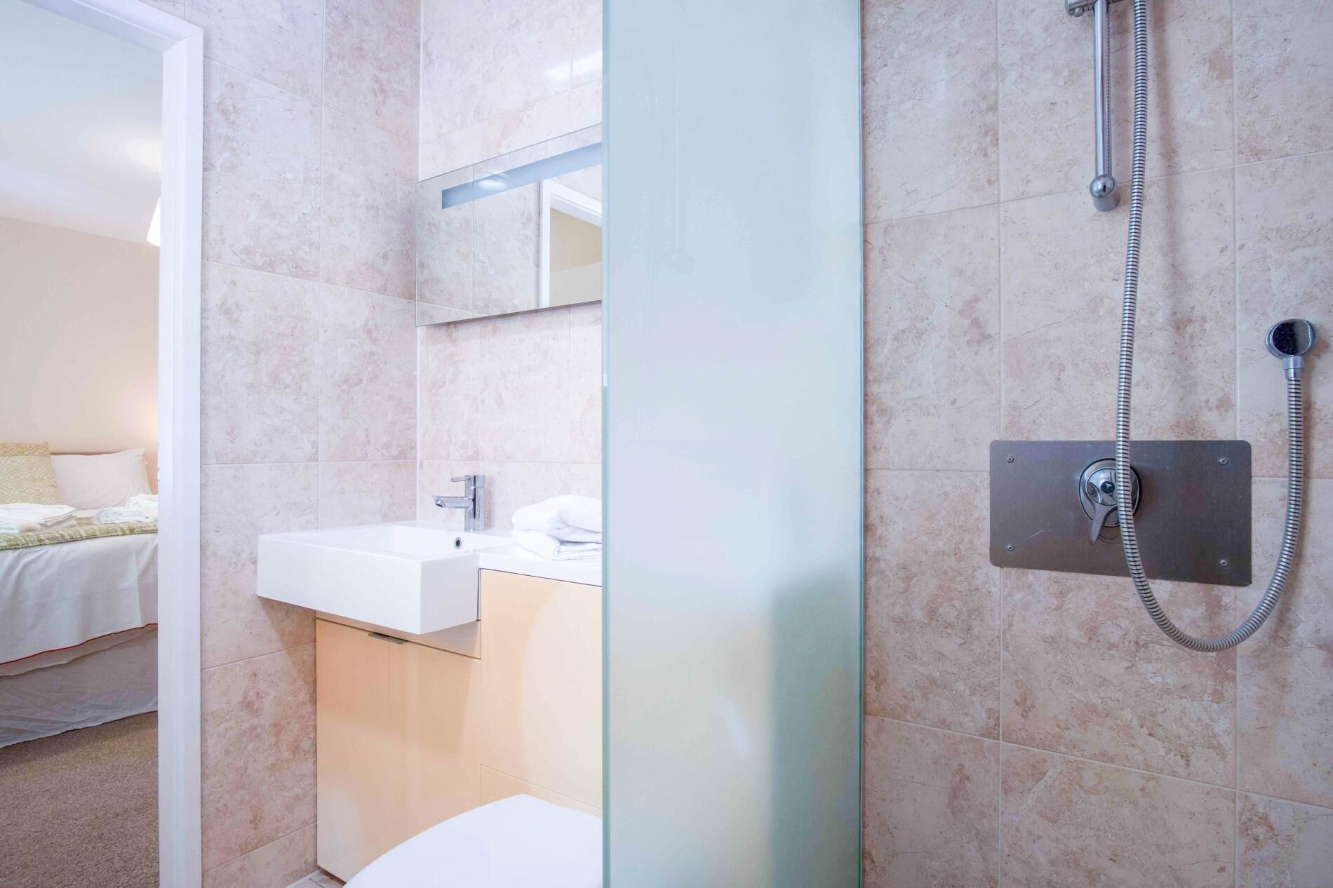 Een moderne badkamer met een douchecabine van glas, een witte wastafel en een bed dat in de aangrenzende kamer zichtbaar is.