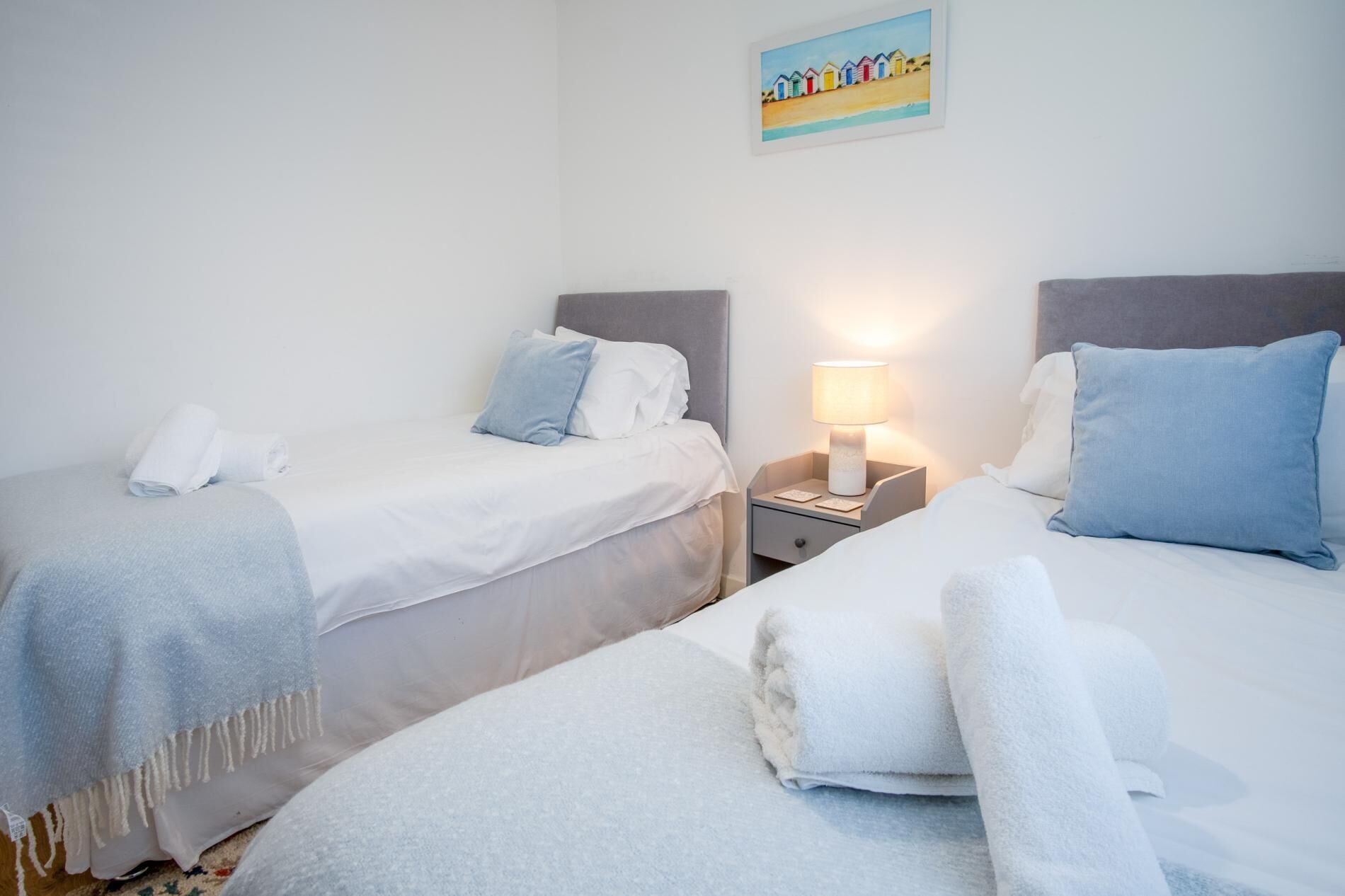 Habitación de hotel con dos camas, cada una con una almohada y una toalla enrollada. Hay una mesita de noche con una lámpara entre las camas.