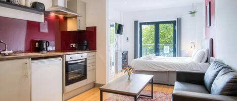 A compact hotel room with a kitchenette, a bed with a view, a sofa, and a small table.