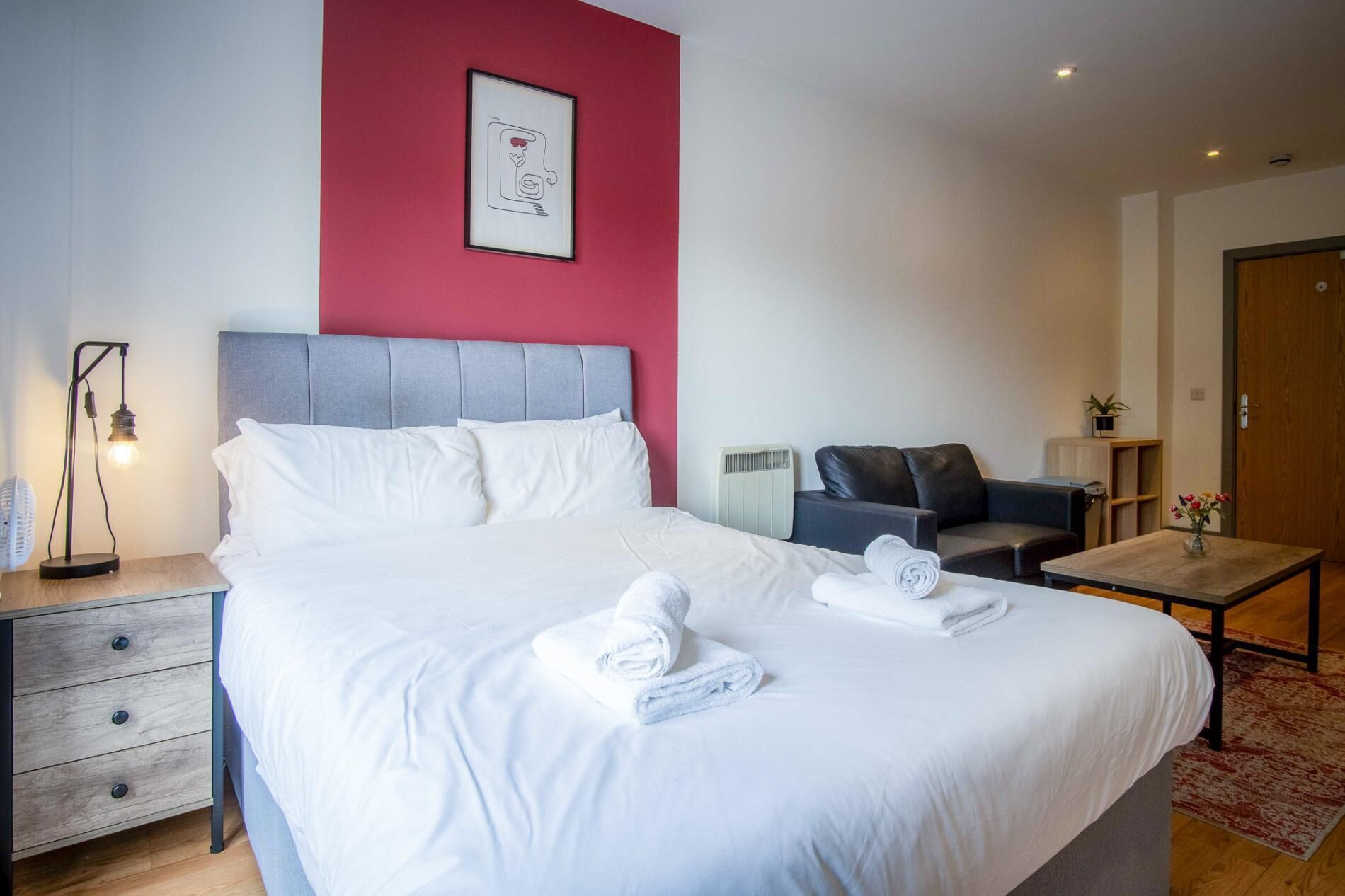 Une chambre d’hôtel avec un grand lit, une table de chevet avec une lampe, un canapé, une petite table avec un vase et une porte.