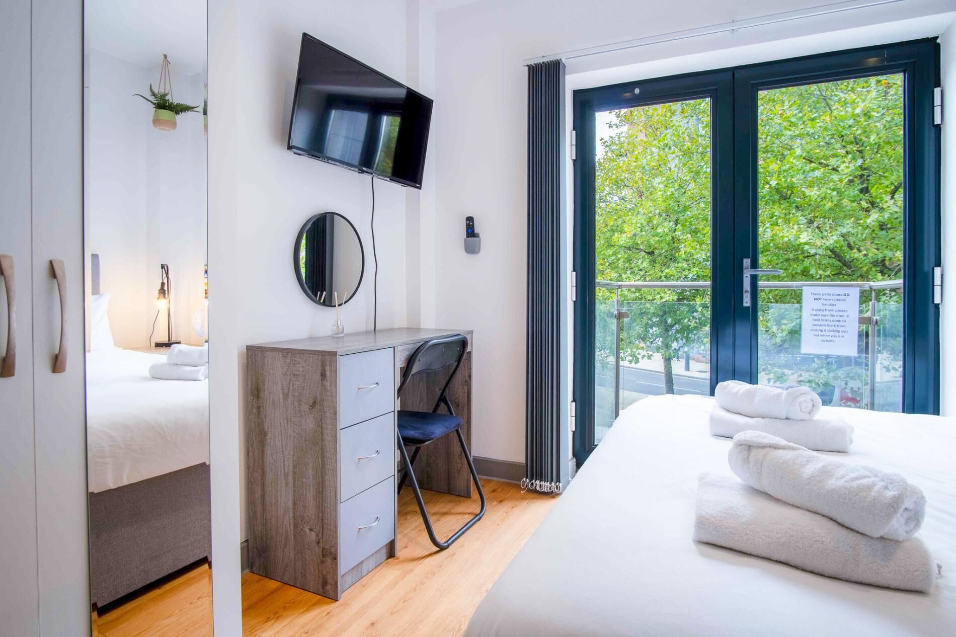 Ein modernes Schlafzimmer mit Bett, Schreibtisch, Fernseher und einem Balkon mit Blick auf Bäume.