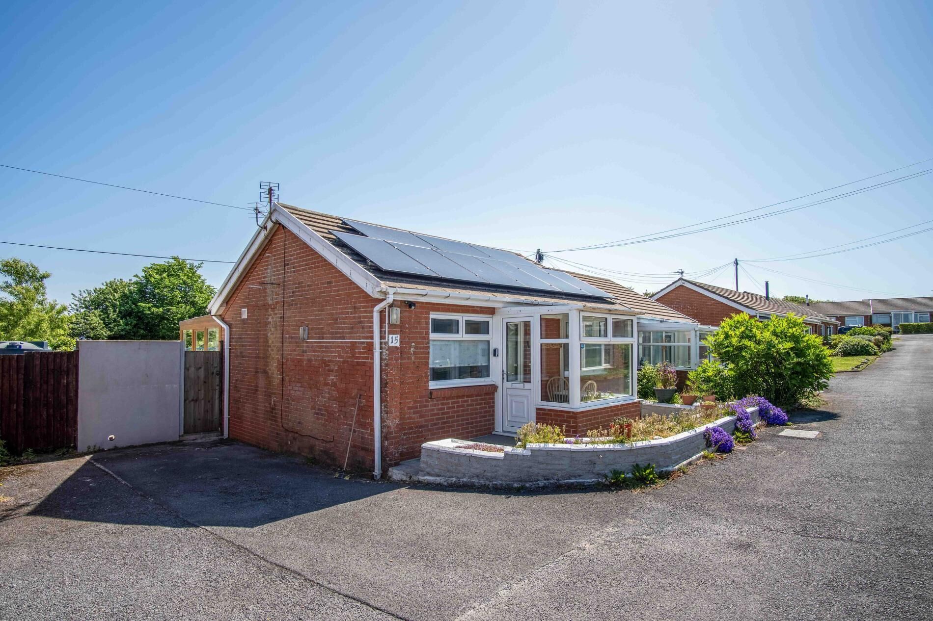 A single-story house with a solar panel roof, a conservatory, and a small garden with purple flowers.