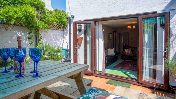 Une terrasse avec une table en bois et de la verrerie bleue, une porte-fenêtre coulissante donnant sur un salon équipé d’un canapé et d’une lampe.