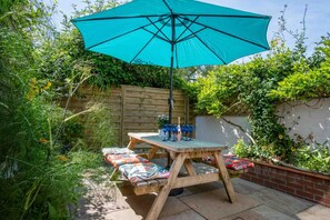 A wooden table with a blue umbrella, surrounded by colorful cushions, in a garden setting.
