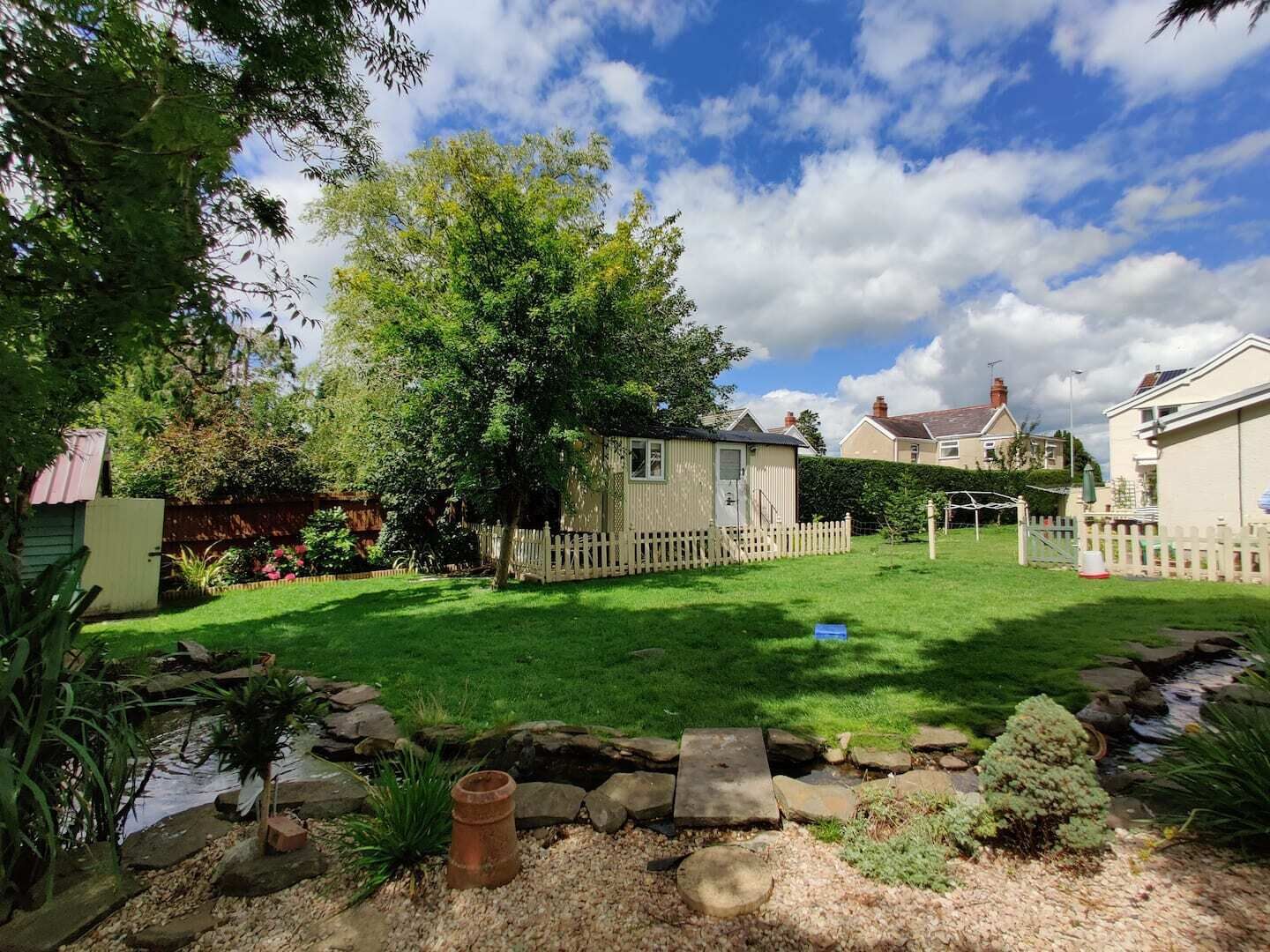 A residential backyard with a well-maintained lawn, a small shed, a wooden fence, and a stone pathway leading to a pond.