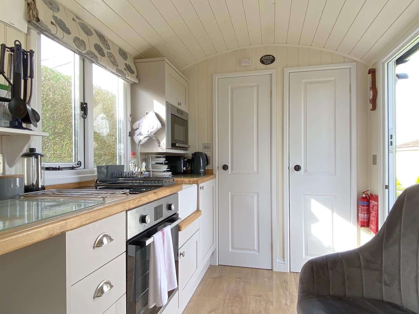 A compact kitchen with a microwave, stove, and sink. There are two white doors and a window with a view of greenery.