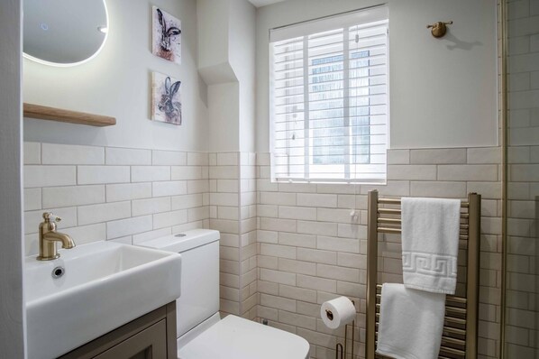 A bathroom with white tiles, a sink, a toilet, a window with blinds, and a towel rack. - Wood Cottage - 3 Bedroom - Bishopston Swansea (Swansea)